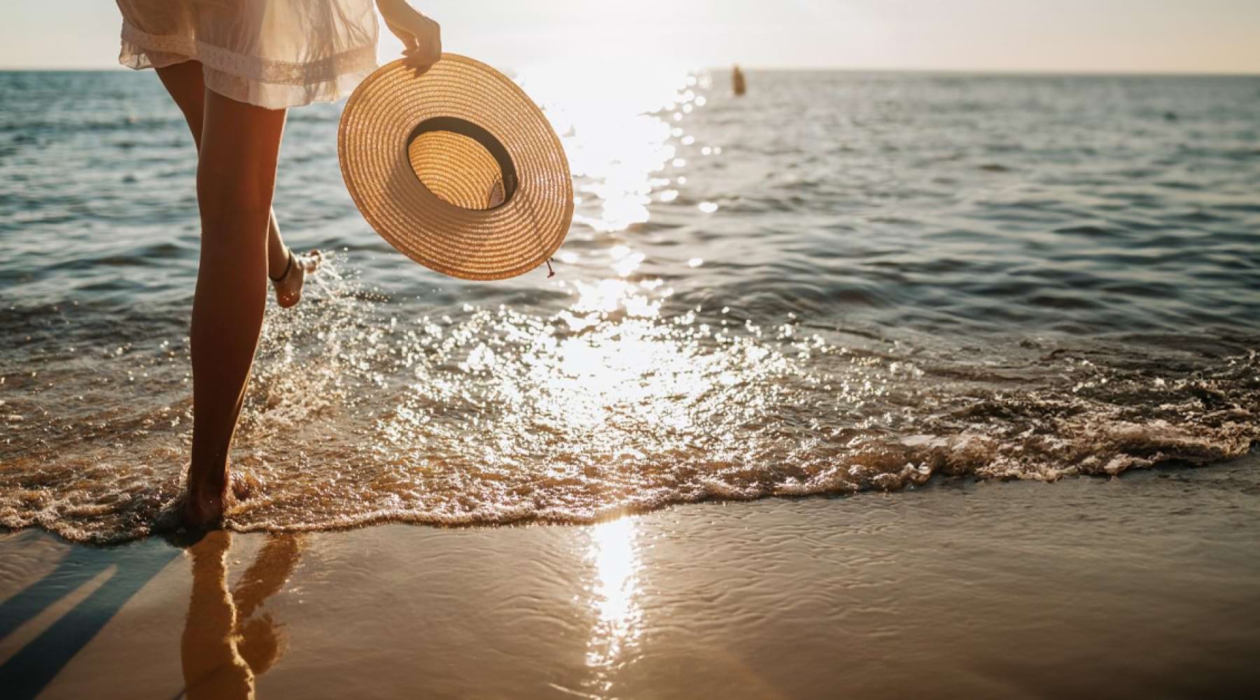 a woman walking into the water of a beach while holding her sunhat at sunset