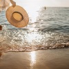 a woman walking into the water of a beach while holding her sunhat at sunset